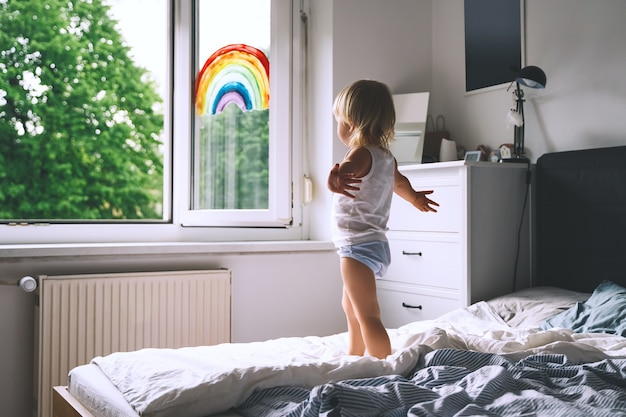 Menina bonitinha se divertindo pulando na cama no fundo da janela com um arco-íris pintado