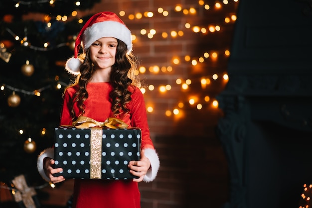 Menina bonitinha segurando um presente perto da árvore de Natal com uma fantasia de Papai Noel vermelho