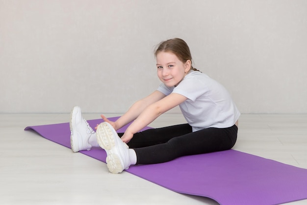 Menina bonitinha senta-se em um tapete de ginástica e faz exercícios de alongamento puxa as mãos para os pés