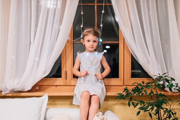 Menina bonitinha senta-se no quarto em um peitoril da janela à noite.