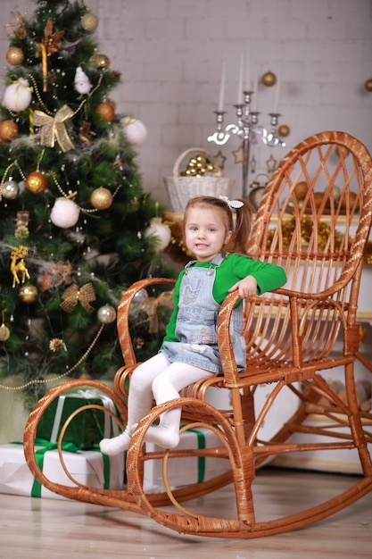 Menina bonitinha sentada na cadeira de balanço perto da árvore de Natal.