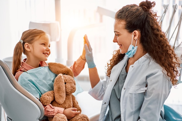 Foto menina bonitinha sentada na cadeira do dentista dando mais cinco para a médica e rindo assistência odontológica confiança e atendimento ao paciente odontologia infantilluz solar