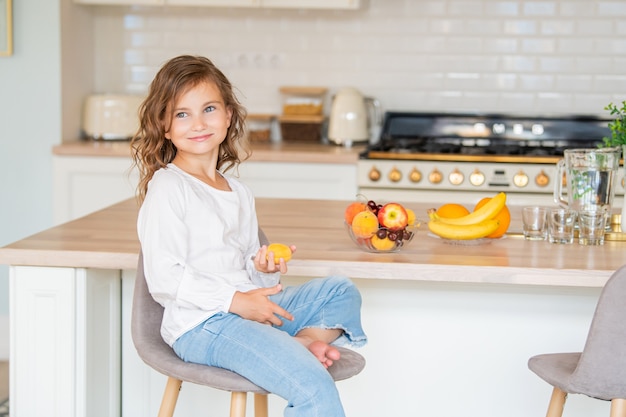 Menina bonitinha sentada na cozinha perto da mesa com frutas e segurando um damasco nas mãos