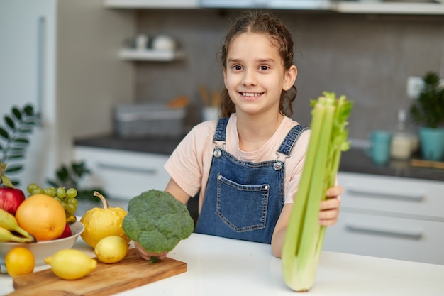 Menina bonitinha sorridente fica perto da mesa na cozinha, segurando brócolis verde e aipo nas mãos.