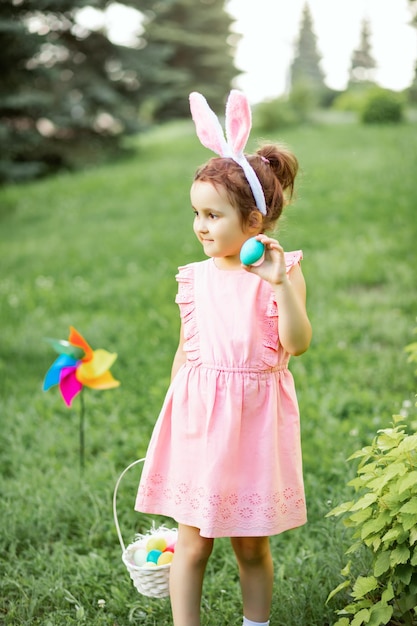 Menina bonitinha usa orelhas de coelho segurando cesta com ovos pintados coloridos na caça aos ovos de Páscoa no parque