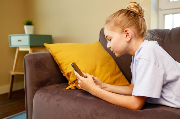 Menina bonitinha usando smartphone e deitada no sofá na sala de estar em sua casa