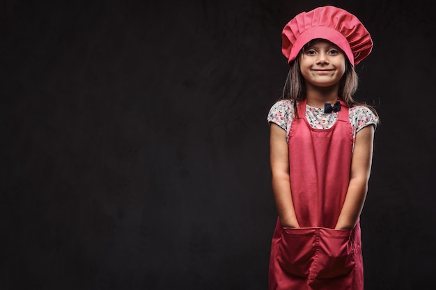 Menina bonitinha vestida com uniforme de cozinheiro rosa segura os braços nos bolsos. Isolado em um plano de fundo texturizado escuro.