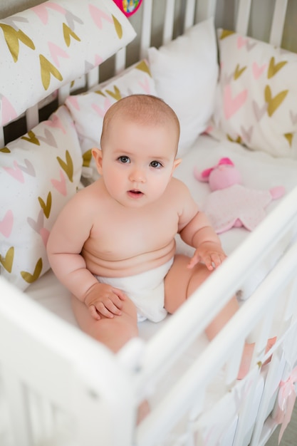 Menina bonito 8 meses senta-se na cama de bebê em casa