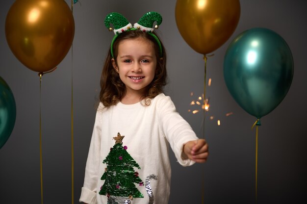 Menina bonito com arco de duende e camisola com árvore de Natal detém estrelinhas e sorrisos contra a decoração de fundo cinza com balões de ar brilhantes verdes dourados de luxo. Criança adorável na festa de ano novo