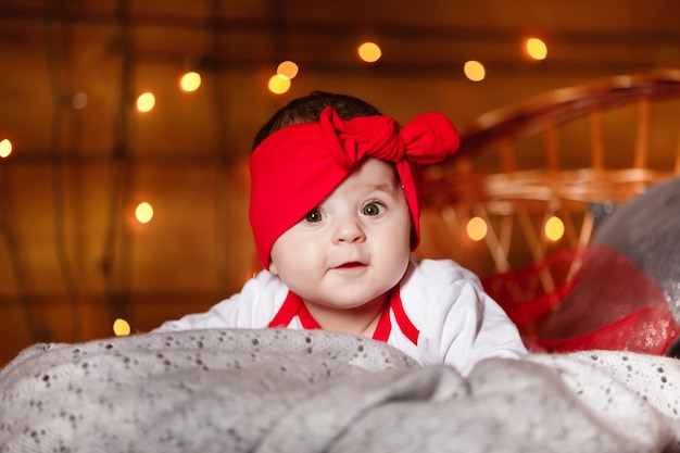 Menina bonito com laço vermelho na cabeça e suéter branco deitado no cobertor, um plano de fundo de Natal em um estúdio com cenário de Natal.