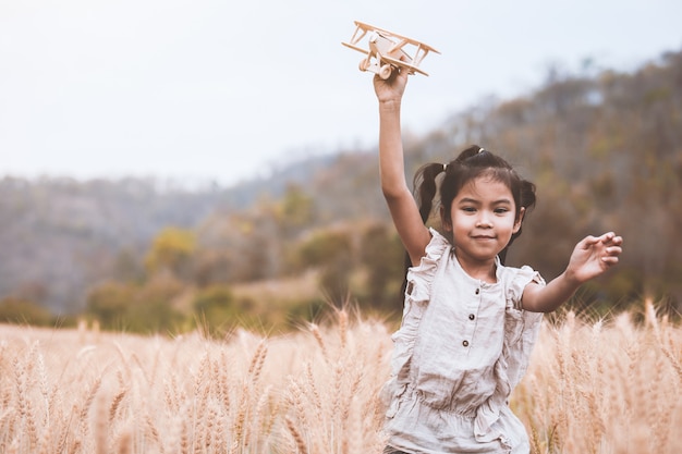 Menina bonito criança asiática correndo e brincando com o avião de brinquedo de madeira no campo de cevada