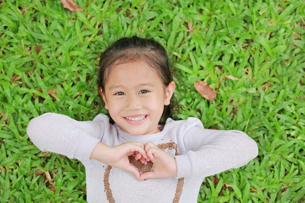 Menina bonito de sorriso que encontra-se no gramado verde com fazer suas mãos para o sinal do coração