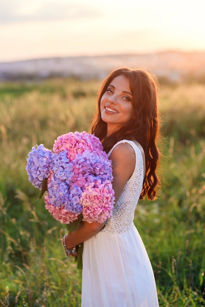 Menina bonito sensual em um vestido branco com um buquê de flores coloridas macias ao pôr do sol, close-up.