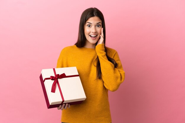 Menina brasileira adolescente segurando um presente sobre um fundo rosa isolado com expressão facial surpresa e chocada