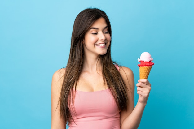 Menina brasileira adolescente segurando um sorvete de corneta sobre fundo azul isolado com expressão feliz