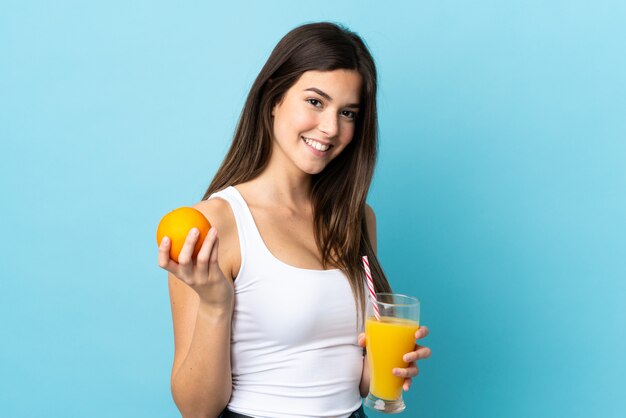 Menina brasileira adolescente sobre parede azul isolada, segurando uma laranja e um suco de laranja