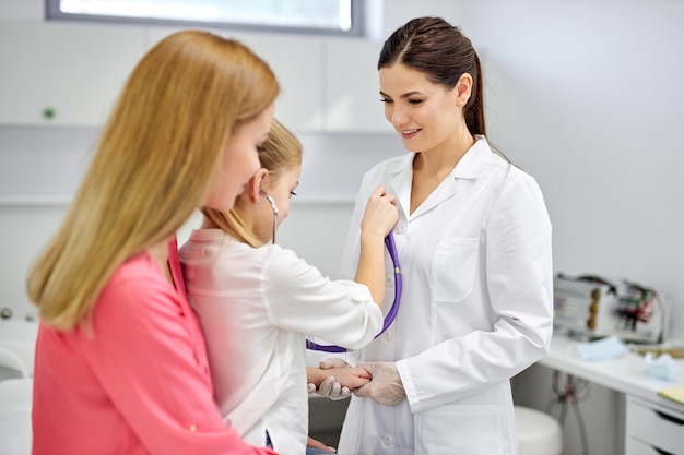 Menina brincando com estetoscópio no hospital, divirta-se com a mulher médica em traje médico, ouça o batimento cardíaco da enfermeira. medicina, conceito de saúde