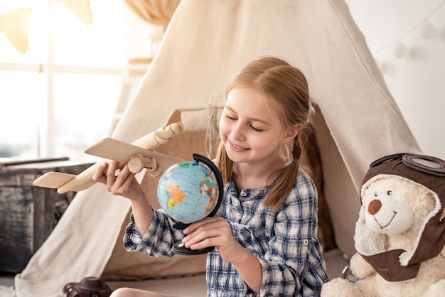 Menina brincando com um globo e um avião de madeira sentada perto da cabana no quarto das crianças