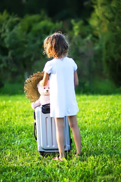 Menina brincando com urso de brinquedo sentado na mala de viagem no prado verde de verão.