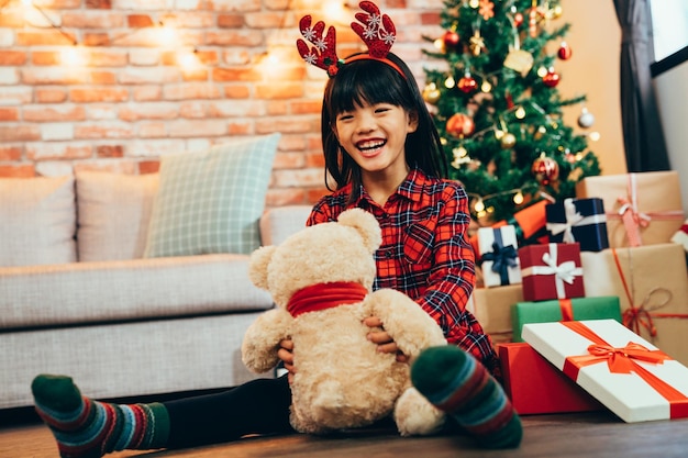 menina brincando de ursinho se divertindo sentado no chão. infância alegre recebeu presente de Natal na celebração do dia do boxe em casa. embalagem presente sob a árvore de natal decorada no fundo.