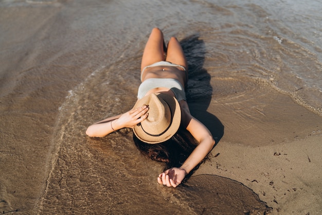 Menina bronzeada em trajes de banho deitado na praia, perto do mar com o chapéu na cara dela.