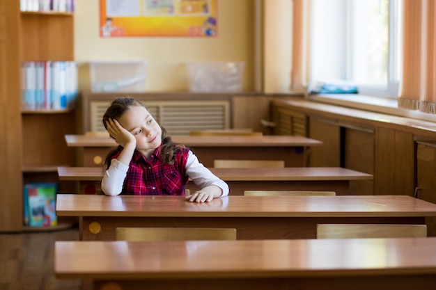 Menina cansado de sorriso que senta-se na mesa na sala de aula e que olha na janela.