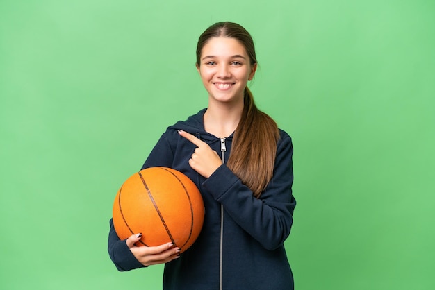 Menina caucasiana adolescente jogando basquete sobre fundo isolado, apontando para o lado para apresentar um produto