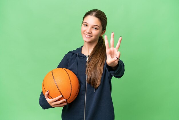 Menina caucasiana adolescente jogando basquete sobre fundo isolado feliz e contando três com os dedos