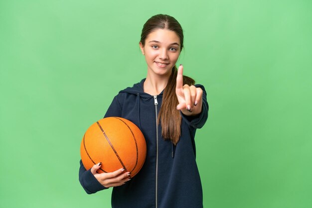 Menina caucasiana adolescente jogando basquete sobre fundo isolado mostrando e levantando um dedo