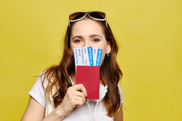 Menina caucasiana cobre metade do rosto com passagens aéreas e passaporte em um fundo amarelo.