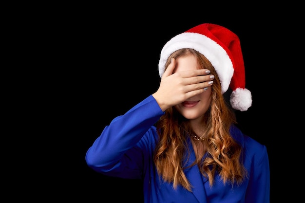 Menina caucasiana com cabelo comprido em um vestido azul com um chapéu de Papai Noel cobre o rosto com a palma da mão isolada em um fundo preto