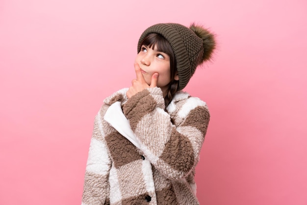 Menina caucasiana com jaqueta de inverno isolada em fundo rosa com dúvidas e com expressão de rosto confuso