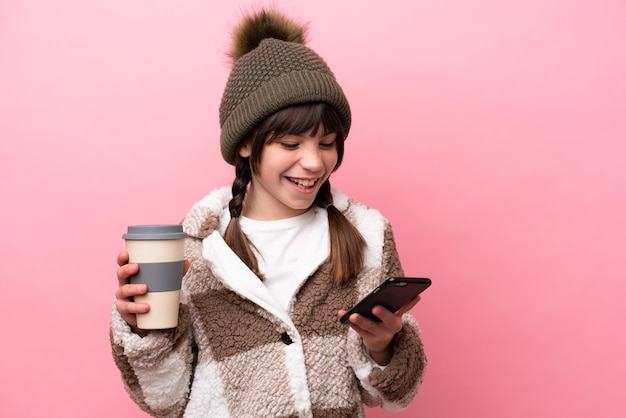 Menina caucasiana com jaqueta de inverno isolada em fundo rosa segurando café para levar e um celular