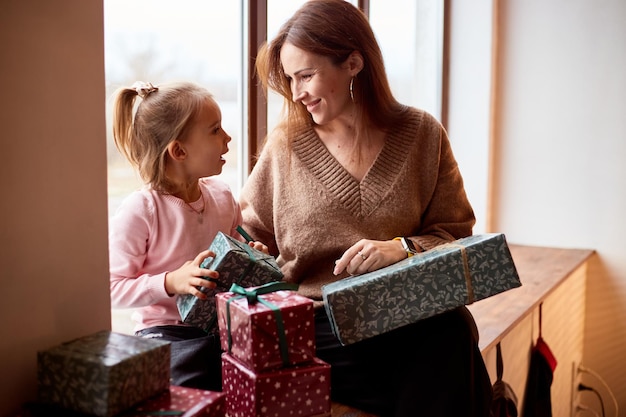 Menina caucasiana com mãe desembalando caixas de presente em casa perto da janela conceito de natal em família feliz