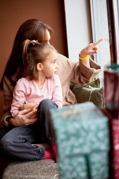 Menina caucasiana com mãe olhando para a janela e sonhando Muitas caixas de presente em torno do conceito de natal em família feliz