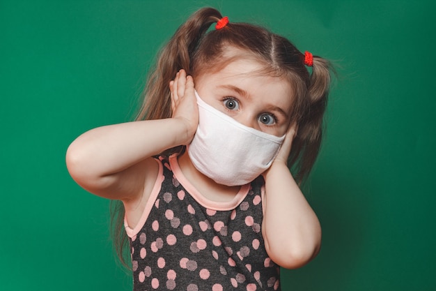 Menina caucasiana com máscara médica usa vestido de bolinhas vermelhas em estúdio sobre fundo verde e segura a cabeça com dor.