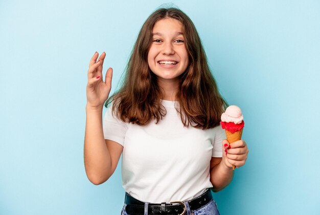 Foto menina caucasiana comendo um sorvete isolado em um fundo azul, recebendo uma agradável surpresa, animada e levantando as mãos.