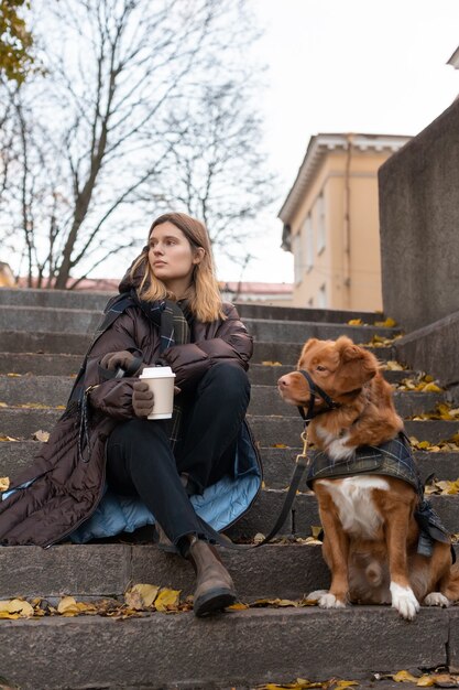 Menina caucasiana de beleza com casaco marrom e com uma xícara de café sentado com o nova scotia duck tolling retriever em degraus de pedra. lazer ao ar livre com cachorro. foco seletivo. conceito de amor de animais de estimação.