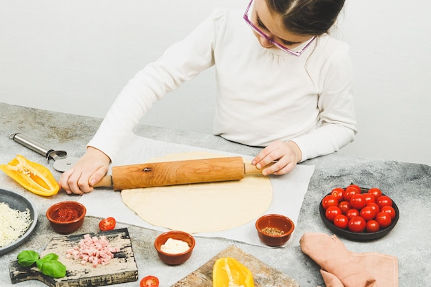 Menina caucasiana de gola alta branca com óculos desenrola a massa de pizza com um rolo