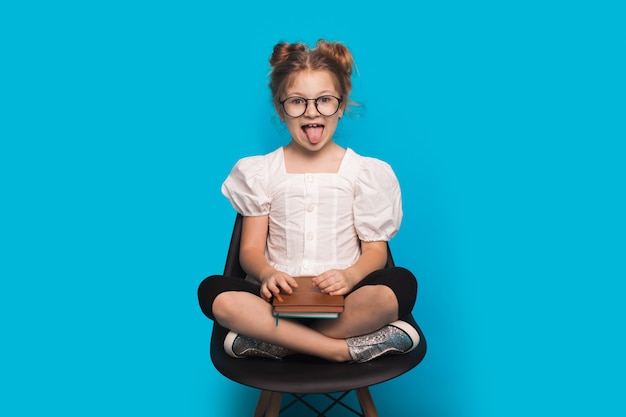 Menina caucasiana de óculos está sorrindo e mostrando a língua enquanto está sentada na poltrona segurando um livro na parede azul do estúdio