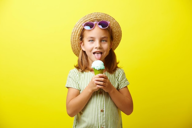 Menina caucasiana de vestido verde e chapéu de palha comendo sorvete lindo usa óculos de sol azuis tem um bom humor fica em fundo amarelo isolado