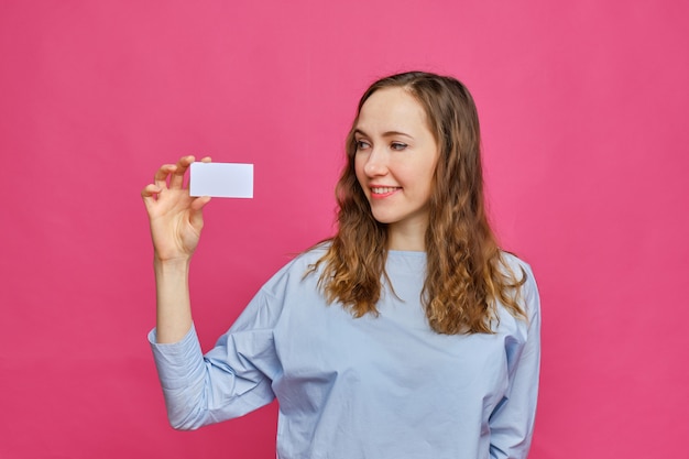 Menina caucasiana elegante em uma camiseta azul pálida tem um cartão branco nas mãos e olha para ele em um fundo rosa.