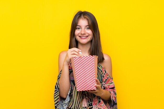 Menina caucasiana em vestido colorido isolado parede amarela comendo pipocas