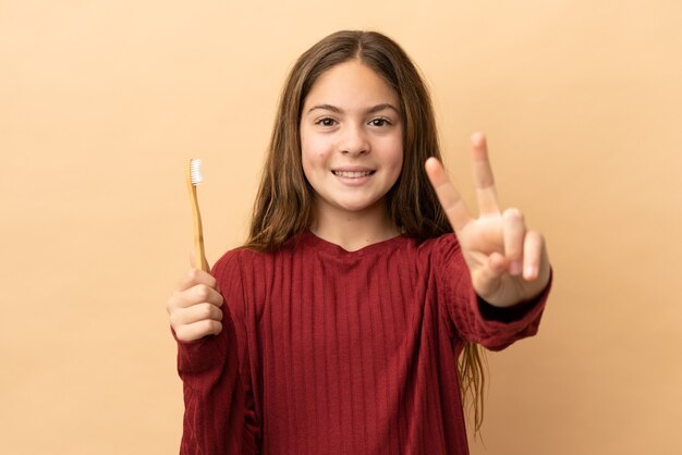 Menina caucasiana, escovando os dentes isolados em um fundo bege, sorrindo e mostrando sinal de vitória