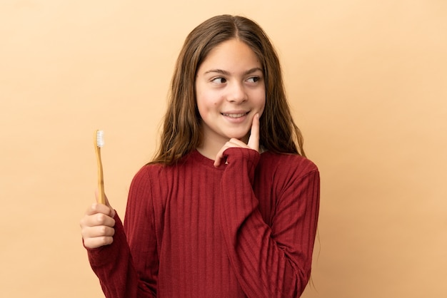 Menina caucasiana, escovando os dentes isolados em um fundo bege, tendo uma ideia enquanto olha para cima