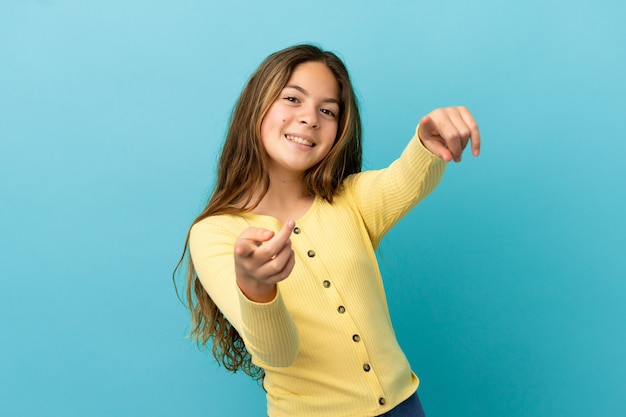Menina caucasiana isolada em um fundo azul apontando para a frente com uma expressão feliz