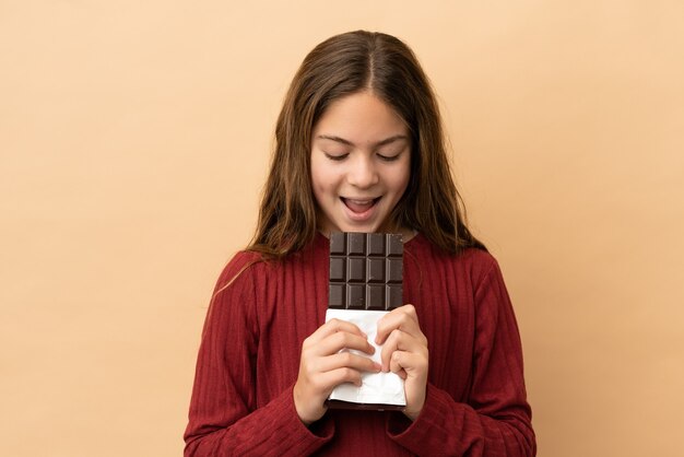 Menina caucasiana isolada em um fundo bege comendo uma tablete de chocolate