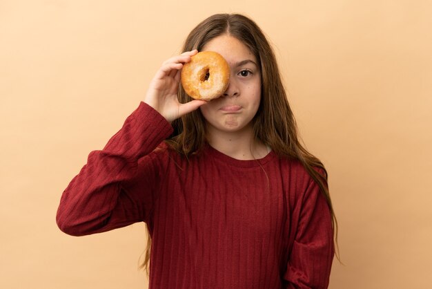 Menina caucasiana isolada em um fundo bege segurando rosquinhas nos olhos com uma expressão triste