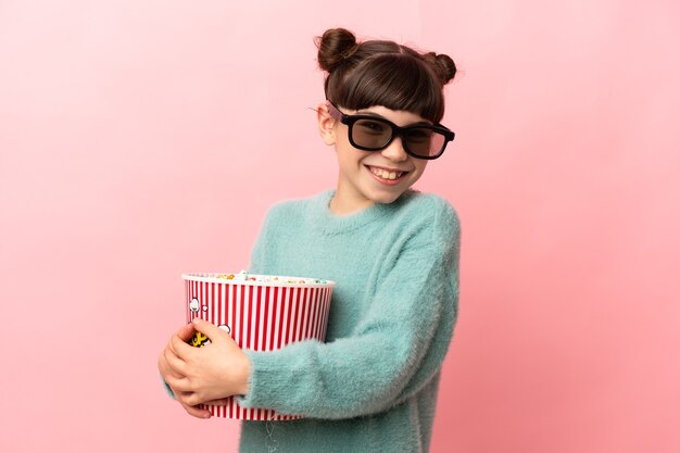 Menina caucasiana isolada em um fundo rosa com óculos 3d e segurando um grande balde de pipocas