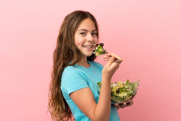 Menina caucasiana isolada em um fundo rosa segurando uma tigela de salada com uma expressão feliz